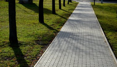 Birbirine kenetlenmiş asfalttan yapılmış gri yol ağaçlardan oluşan bir ara sokak ve yeşil çimlerle kaplı. Yol doğrudan mesafeye çıkıyor.