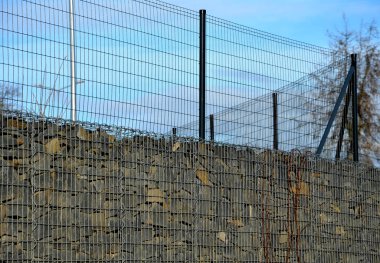 Gabon sepetlerinden yapılmış istinat duvarı. Yüksek gri bir duvarın tepesinde hala bir tel var. Duvarın altında arabalar için beton bir otopark var.