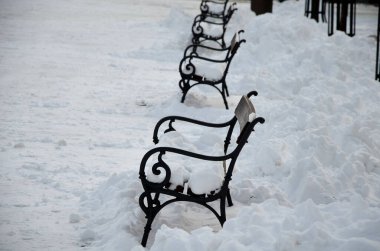 Metal bankların olduğu bir kare. Meydan karla kaplı ve traktörün kenara ittiği yığınları görebilirsiniz. Banklar büyük bir kar tabakasıyla kaplı. Kimse oturmuyor ve...