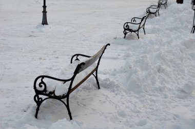 Metal bankların olduğu bir kare. Meydan karla kaplı ve traktörün kenara ittiği yığınları görebilirsiniz. Banklar büyük bir kar tabakasıyla kaplı. Kimse oturmuyor ve...