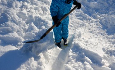 Çocuk evin etrafında bir kar felaketini temizlemeye çalışıyor. Elinde bir kürek, bir kürek tutuyor ve mavi bir tulumla karda çalışıyor..