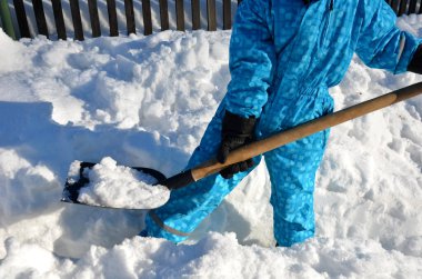 Çocuk evin etrafında bir kar felaketini temizlemeye çalışıyor. Elinde bir kürek, bir kürek tutuyor ve mavi bir tulumla karda çalışıyor..