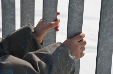 Metal kafes parmaklığı tutan bir çocuk. Elleri diğer tarafa geçmeyi deniyor. Dairenin balkonundan düşme tehlikesi. Merdivenlerde güvenlik koruması