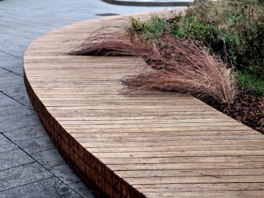 Çember şeklinde büyük ahşap bir bank, tepenin altında bir çiçek yatağı ve çimen var.