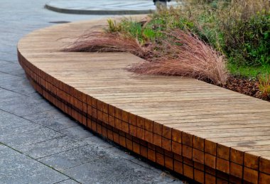 Çember şeklinde büyük ahşap bir bank, tepenin altında bir çiçek yatağı ve çimen var.