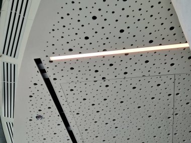 Delikli alçı kaplama tavan panelleri fayanslı zemini olan büyük bir salonda yankılanıyor. Seslerin boğuk, baskının, ses sisteminin, ahşap zeminin duyulduğu bir otele giriş salonu.