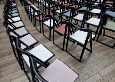 Koltuklarında renkli yastıklar olan siyah plastik katlanır sandalyeler, ahşap bir zeminde sıra sıra dizilmiş. Ödül töreni için hazırlık ya da salonda hazırlıksız konferans.