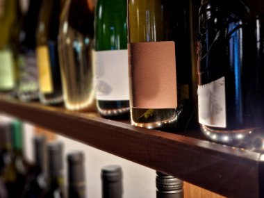 Ahşap raflarda sergilenen şarap şişeleri. Bu, beyaz, gül ve kırmızı dahil olmak üzere farklı türde şarap koleksiyonudur. Şişelerin şekil ve boyutları farklı. LED şeritli arka ışıklandırma