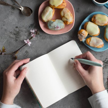 Kadın iki eli boş bir not tutun. Tasarımcısı tablo kahve molası çizimleri çizer. Tükenmez kalem defter üzerinde yazma elle. Kuş gözü görüntüleyin. Pastel tonları. Dal bahar kiraz çiçeği.