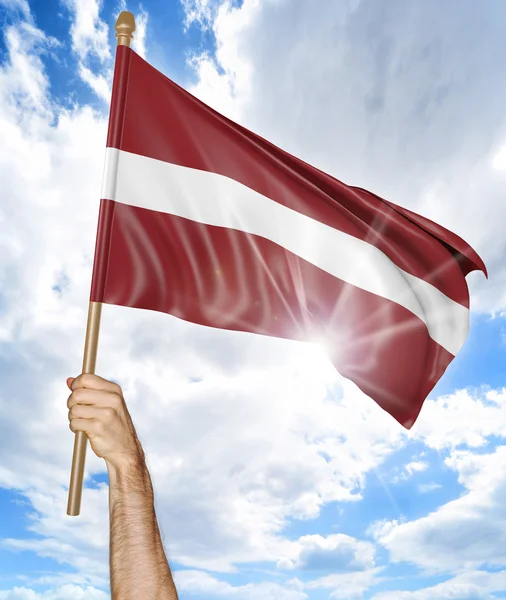 La mano della persona che tiene la bandiera nazionale lettone e la sventola nel cielo, rendering 3D — Foto Stock