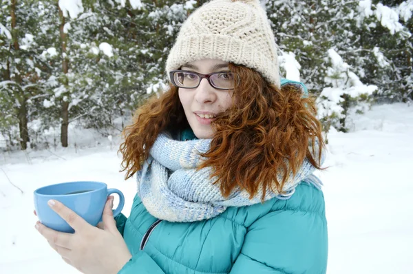 Dark-haired girl dressed in a turquoise jacket,bodily woolly hat, and a light blue scarf,reading a book in the Park,drinking tea,coffee,cocoa