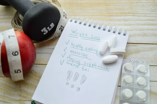 Notebook with pills, apple, measuring tape and dumbbell — Stock Photo, Image