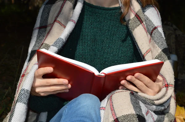 Une jeune et belle fille enveloppée dans une couverture chaude à carreaux buvant du thé chaud et lisant un livre dans le parc — Photo