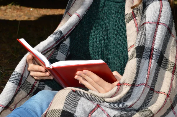 Una joven y hermosa niña envuelta en una cálida manta a cuadros bebiendo té caliente y leyendo un libro en el Parque —  Fotos de Stock