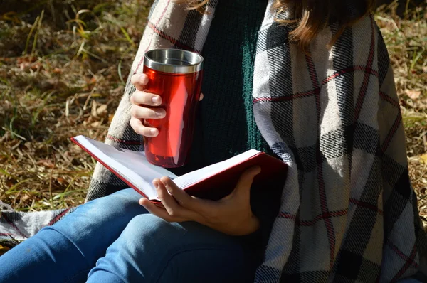 Молода красива дівчина, загорнута в теплу ковдру, п'є гарячий чай і читає книгу в парку — стокове фото