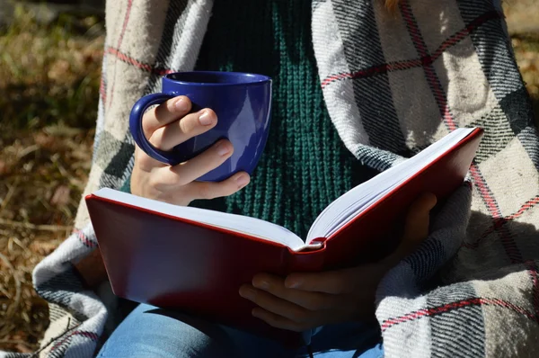 Una joven y hermosa niña envuelta en una cálida manta a cuadros bebiendo té caliente y leyendo un libro en el Parque — Foto de Stock