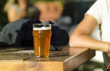 Genç bir adam bir bardak sarı birayla masada oturuyor, açık hava bar sahnesi.