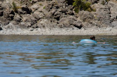 Açık mavi şişirilebilir can yeleği olan çocuk gölde taş zeminde.