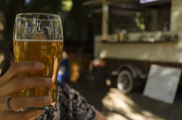 toast with pint of beer, woman\'s hand, selective focus with food truck in background. outdoor craft beer brewery yard