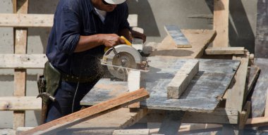 İnşaat işçisi, iş kıyafetleri ve aletlerle testere ile kesiyor. Mavi tulumlar, sarı miğfer, aletleri güvenli bir şekilde kullanabilmek için gözlük.