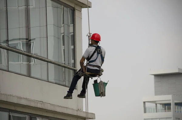 带头盔和桶的高楼玻璃清洁工 用绳子和脚手架吊着 危险工作 — 图库照片