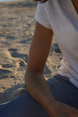 Kadın kumda oturmuş düşünüyor. Kot pantolon ve beyaz gömlek giymişti. Kumsaldaki genç kadın.