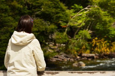 Arkasından beyaz ceketli bir kadın orman ve nehirle kaplı bir köprüdeki manzaraya bakıyor.