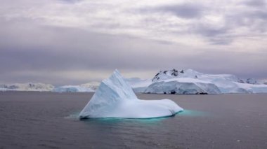 Okyanusta açık mavi bir buz kütlesi var. Arka planda uzun bir buzdağı var..