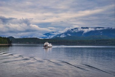 BC, Canada - June 24, 2019: The Ferryboat sailing through the lake, carrying passengers and cars. Beautiful scenery on both sides of the fjord, with layers of mountains and clouds. The view seen from the cruise ship.