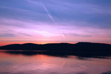 Pembe gökyüzünde beyaz bir çizgi çizilir. Gölün yansıması rengarenk. Alaska fiyortları, eşsiz doğal manzaralar. Alaska, ABD. Haziran 2019.