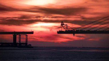 Vibrant sunset with sun partially obscured by clouds over the Tamkang Bridge construction site in Tamsui New Taipei Taiwan. Boat passes under the red and orange sky.