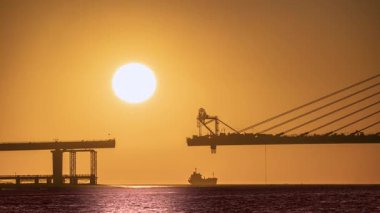 Golden hour sunset over the Tamkang Bridge construction in Tamsui New Taipei Taiwan. A ship passes under the huge setting sun, creating a dramatic silhouette scene.