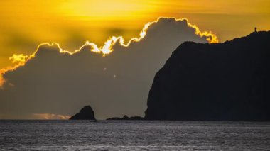 Close-up of the sun rising over the ocean near Wanli New Taipei Taiwan. Golden sky and clouds silhouette the coastal mountain and Xiao Jilong Islet in a dramatic scene.