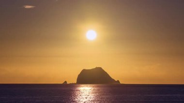 Close-up of the sun rising over the ocean near Wanli New Taipei Taiwan. Golden sky and clouds silhouette the coastal mountain and Xiao Jilong Islet in a dramatic scene.