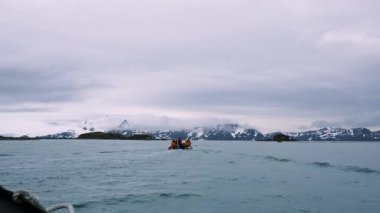 Turuncu can yelekli turistler bir Zodiac teknesinde dramatik karlı dağlara ve bulutlu bir günde Güney Georgia 'da Salisbury Ovası' nın engin manzarasına bakıyorlar..