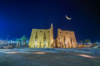 Gece vakti Luxor Tapınağı 'nın aydınlık sütun heykelleri ve dikilitaşının geniş açılı görüntüsü. Göz kamaştırıcı bir hilal ve Venüs gezegeni koyu mavi gökyüzünde görülebilir. 2025 'te çekilmiş..