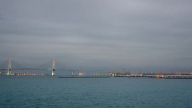 Yokohama Limanı, Japonya 'dan kalkan bir yolcu gemisinden sonbahar alacakaranlık manzarası. Aydınlatılmış Yokohama Körfezi Köprüsü, gemi ufuk boyunca ilerlerken ön planda sakin sularla birlikte uzanıyor..