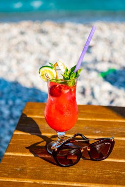 Ahşap bir masada deniz kenarında güneş gözlüklü kırmızı bir kokteyl.