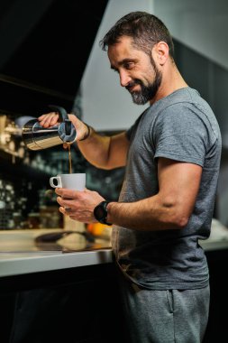                                Genç, siyah saçlı, kahve içen ve evinin mutfağındaki cep telefonuna sıradan kıyafetlerle bakan bir adam.