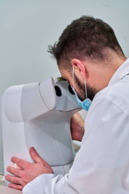 Optometriste çalışan beyaz önlüklü ve optometri makineli bir adam.