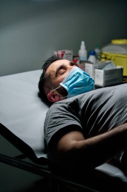 Maskeli beyaz adam sedyede yatıyor ve gözleri kapalı.