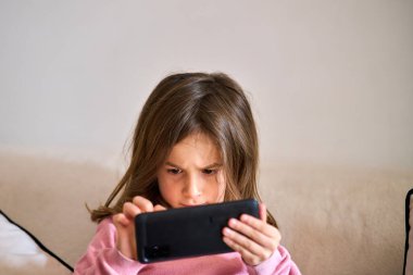 Kanepede oturan küçük bir kız telefonla oynuyor. 