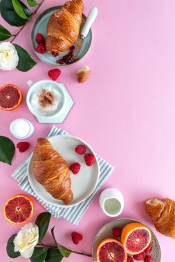 Pembe arka planda kruvasan, kan portakalı, ahududu, çiçek ve kapuçino içeren bir kahvaltı sahnesi.