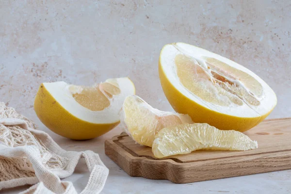A delicious yellow pomelo sliced open with wedges, and a knife on a wooden cutting board with a mesh bag on a neutral background, horizontal with copy space