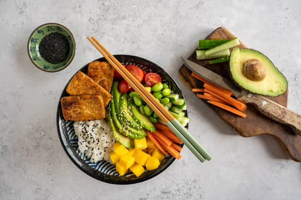 Tofu, mango, avokado, havuç, edamame, domates, salatalık ve gri arka planda kesim tahtasıyla ev yapımı bir vejetaryen dürüm kasesinin en üstten görünüşü.