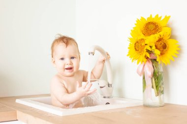 Bebek mutfak lavabosunda banyo yapıyor. Banyo yapan küçük çocuk. Çocuklar için su eğlencesi. Çocuklar için hijyen ve cilt bakımı. Yakınlarda bir buket ayçiçeği duruyor.