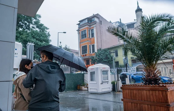 Beyoğlu, İstanbul, Türkiye - 06.18.2021: Siyah şemsiyeli erkek ve kadın aşıklar Taksim bölgesinde yaz mevsiminde sağanak yağmurdan korunmak için binanın yakınında bekliyorlar