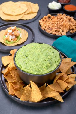 Meksika guacamole 'si ve yanında siyah kase üzerinde tortilla cipsi. Arka planda domuz derisi taco, tortilla, tabakta domuz kabuğu ve salsa sosu gibi Meksika yemekleri var. Portre görünümünde