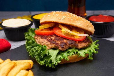 Marullu sığır burger, domates, hardal, soğan halkaları ve patates kızartması ve kahverengi bira ile birlikte taş tabakta turşu. Taş yüzey ve mavi arkaplan fotokopi alanı. Kapat.