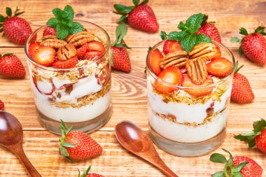 Birkaç bardak yulaf ezmesi, bal ve üzerinde nane yaprağı olan fındıklı çilekli parfe. Bir tarafta bütün çilekler ve tahta kaşıklar, ahşap bir masada..